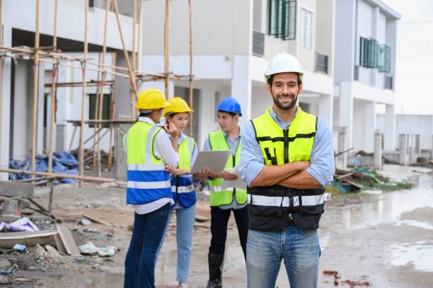 young attractive construction man smiling bearded in vest with white helmet standing on construction site